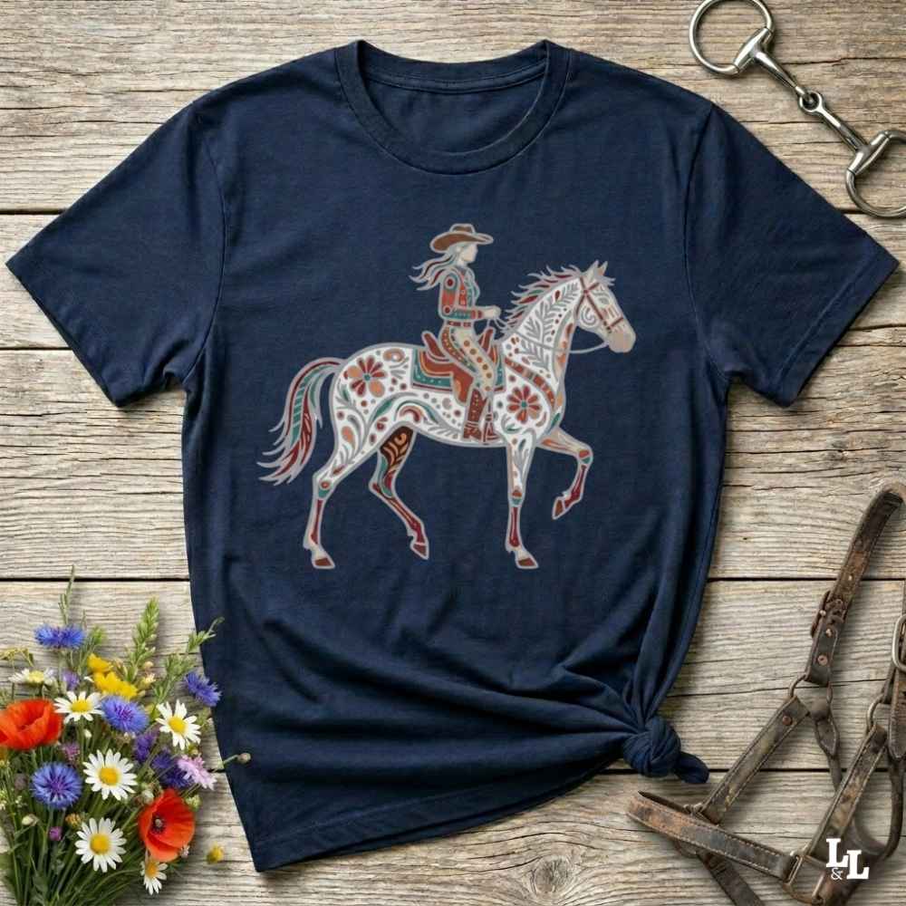 A navy blue short-sleeve t-shirt with a colorful graphic of a cowboy riding a decorated horse, styled on a rustic wooden background with equestrian accessories and flowers.