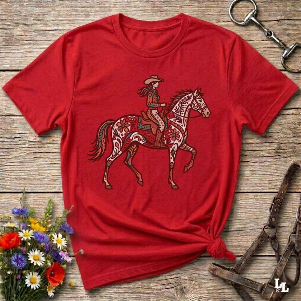 A red short-sleeve graphic t-shirt featuring a cowboy riding a decorated horse, laid flat on a rustic wooden surface with equestrian accessories and flowers around it.