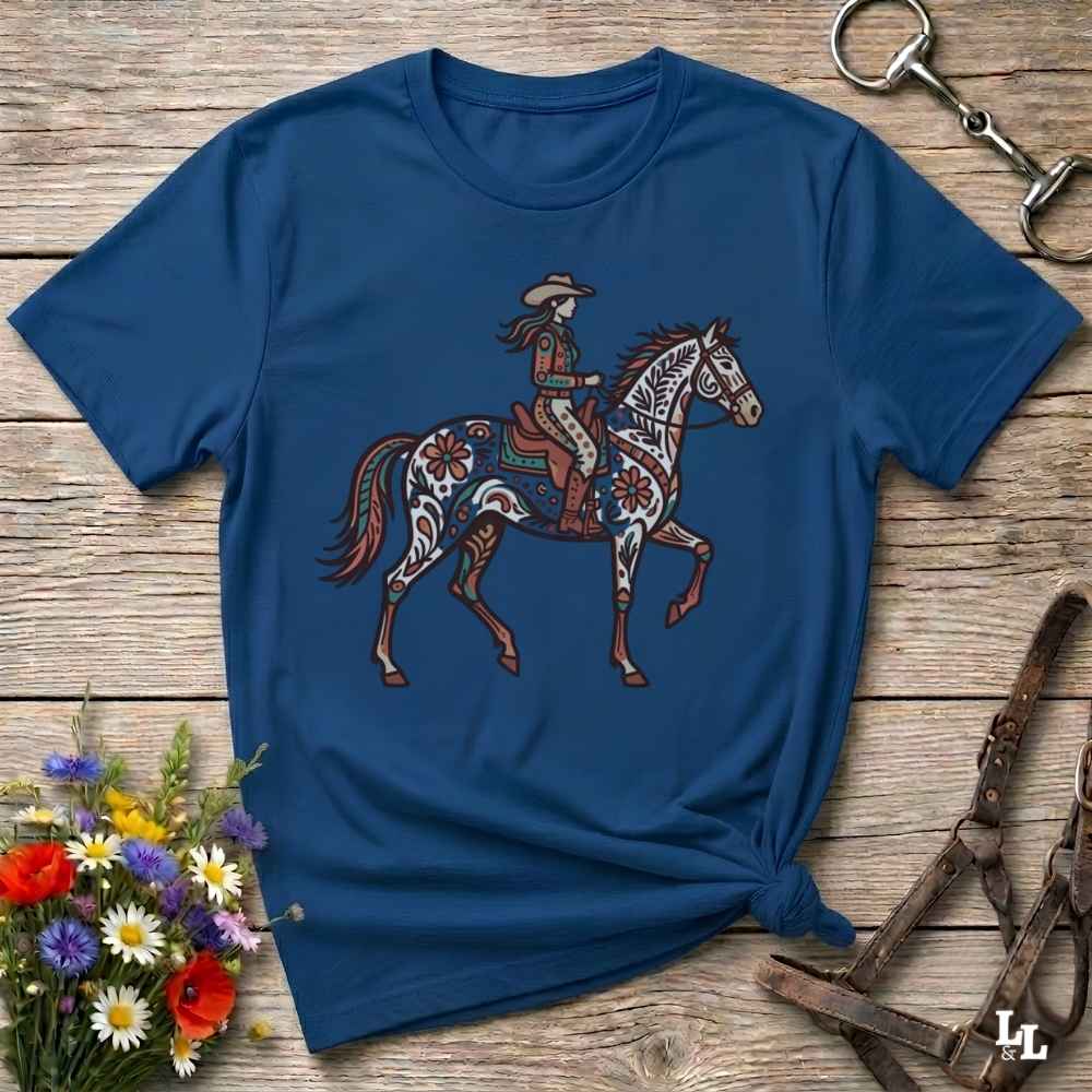 A navy blue short-sleeve t-shirt with a western-themed graphic of a cowboy riding a decorated horse, laid flat on a rustic wooden surface with floral and tack props.