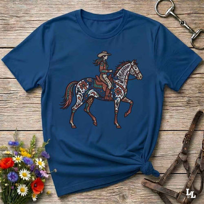 A navy blue short-sleeve t-shirt with a western-themed graphic of a cowboy riding a decorated horse, laid flat on a rustic wooden surface with floral and tack props.