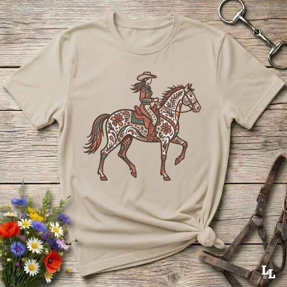 A beige short-sleeve t-shirt with a graphic of a cowboy riding a decorated horse, styled on a rustic wooden background with equestrian accessories and flowers.