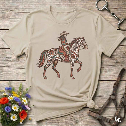 A beige short-sleeve t-shirt with a graphic of a cowboy riding a decorated horse, styled on a rustic wooden background with equestrian accessories and flowers.