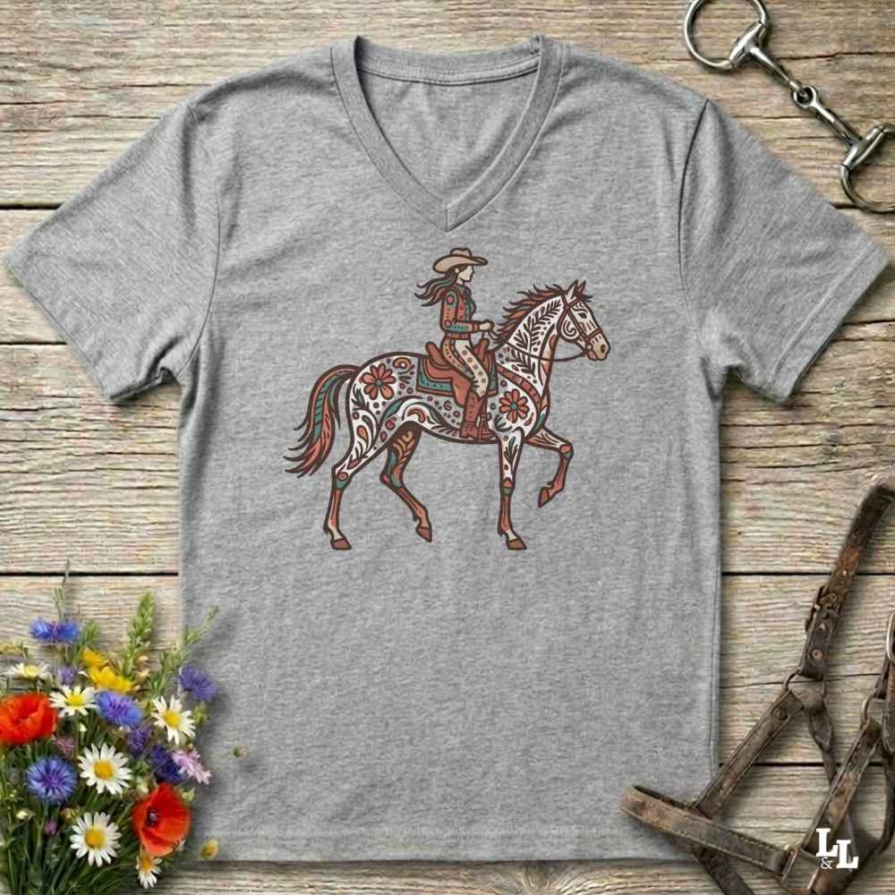 A heather gray V-neck t-shirt featuring a graphic of a cowboy riding a patterned horse, laid flat on a rustic wooden surface with decorative flowers and horse tack props.