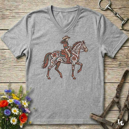 A heather gray V-neck t-shirt featuring a graphic of a cowboy riding a patterned horse, laid flat on a rustic wooden surface with decorative flowers and horse tack props.