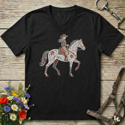A black V-neck t-shirt featuring a western-themed graphic of a cowboy riding a decorated white horse, laid flat on a rustic wooden background with flowers and horse tack props.
