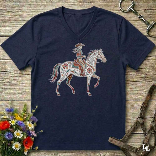 A navy blue V-neck t-shirt featuring a graphic of a cowboy riding a white horse decorated with red and orange patterns, laid flat on a rustic wooden background with equestrian accessories and flowers.