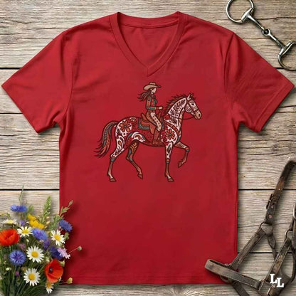 A red V-neck t-shirt featuring a graphic of a cowboy riding a patterned horse, laid flat on a rustic wooden background with equestrian accessories and flowers.