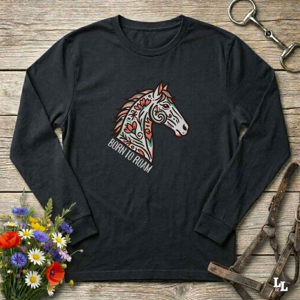 Black long-sleeve shirt with a colorful horse graphic and 'Born to Roam' text on a wooden surface with floral and equestrian-themed accessories.