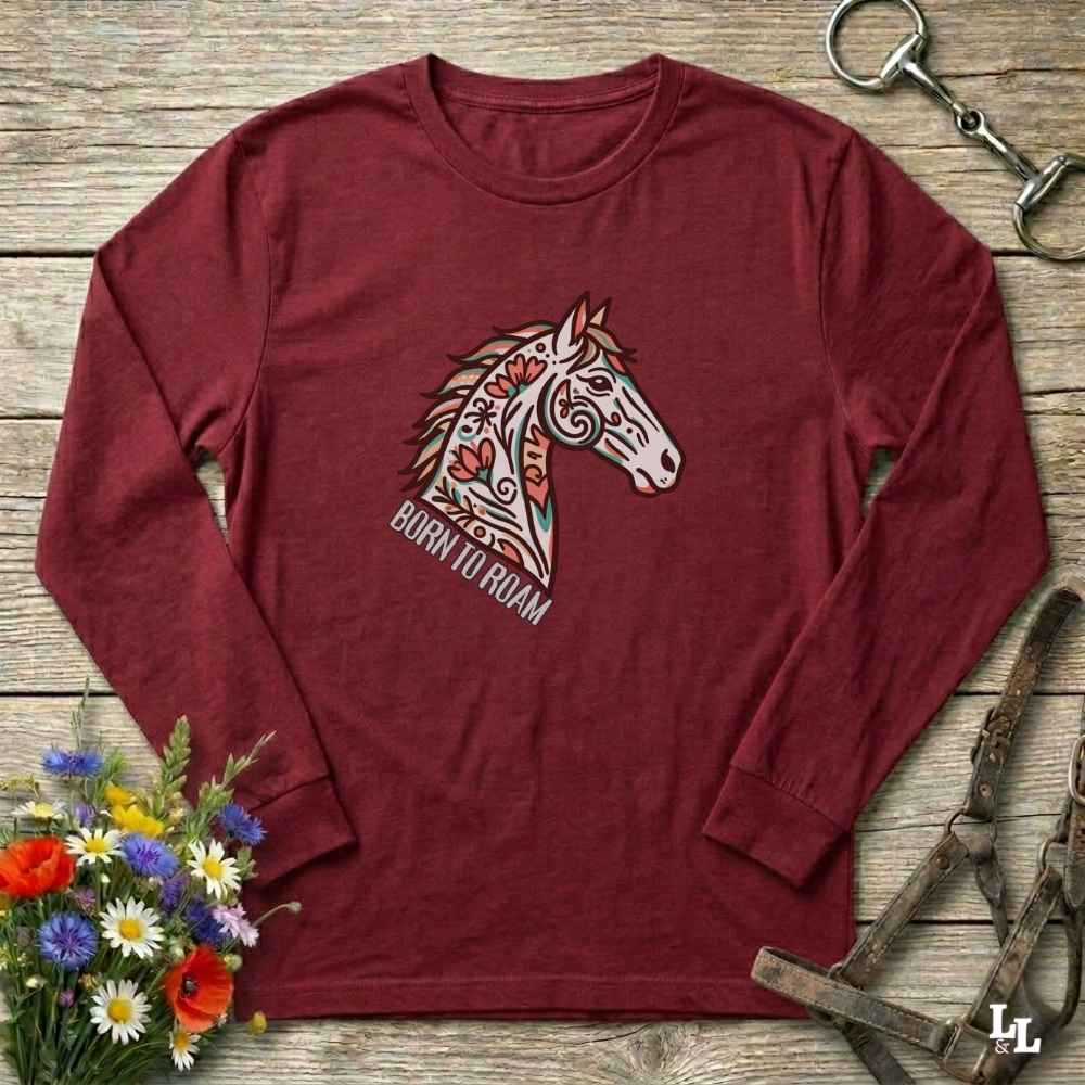 Maroon long-sleeve shirt with horse graphic and 'Born to Roam' text on a wooden surface with flowers and equestrian equipment.