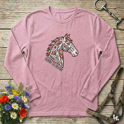 Pink long-sleeve shirt with a horse design and 'Born to Roam' text on a wooden surface with flowers and equestrian equipment.