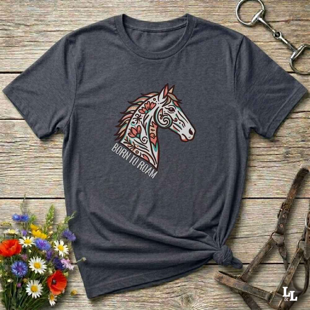 Gray t-shirt with a colorful horse graphic and 'Born to Roam' text on a wooden surface with flowers and equestrian equipment.