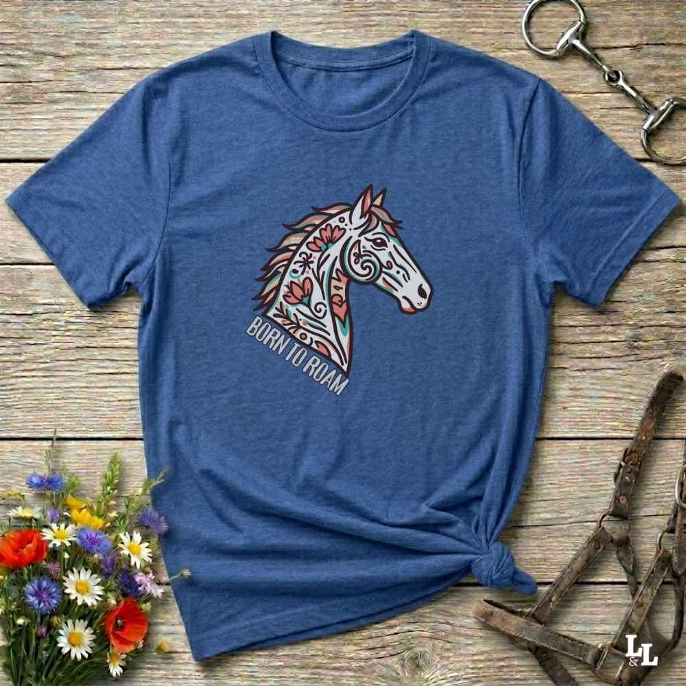 Blue t-shirt with a colorful horse graphic and text on a wooden surface with flowers and equestrian equipment.