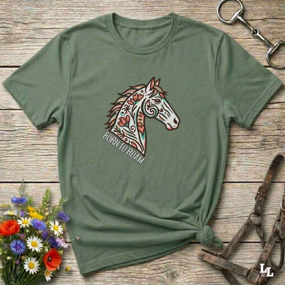 Green t-shirt with a horse graphic and 'Born to Roam' text on a wooden surface with flowers and equestrian equipment.