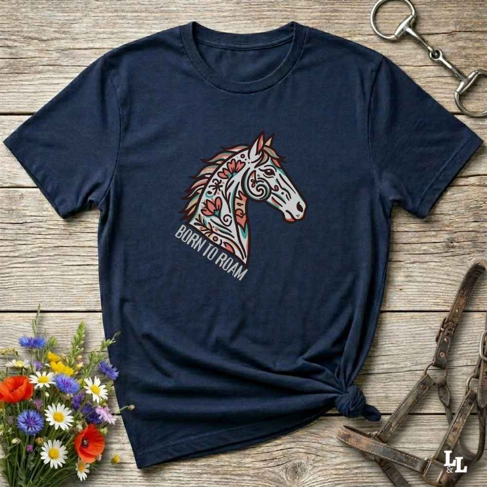 Navy blue t-shirt with a colorful horse graphic and 'Born to Roam' text on a wooden surface with floral and equestrian-themed accessories.