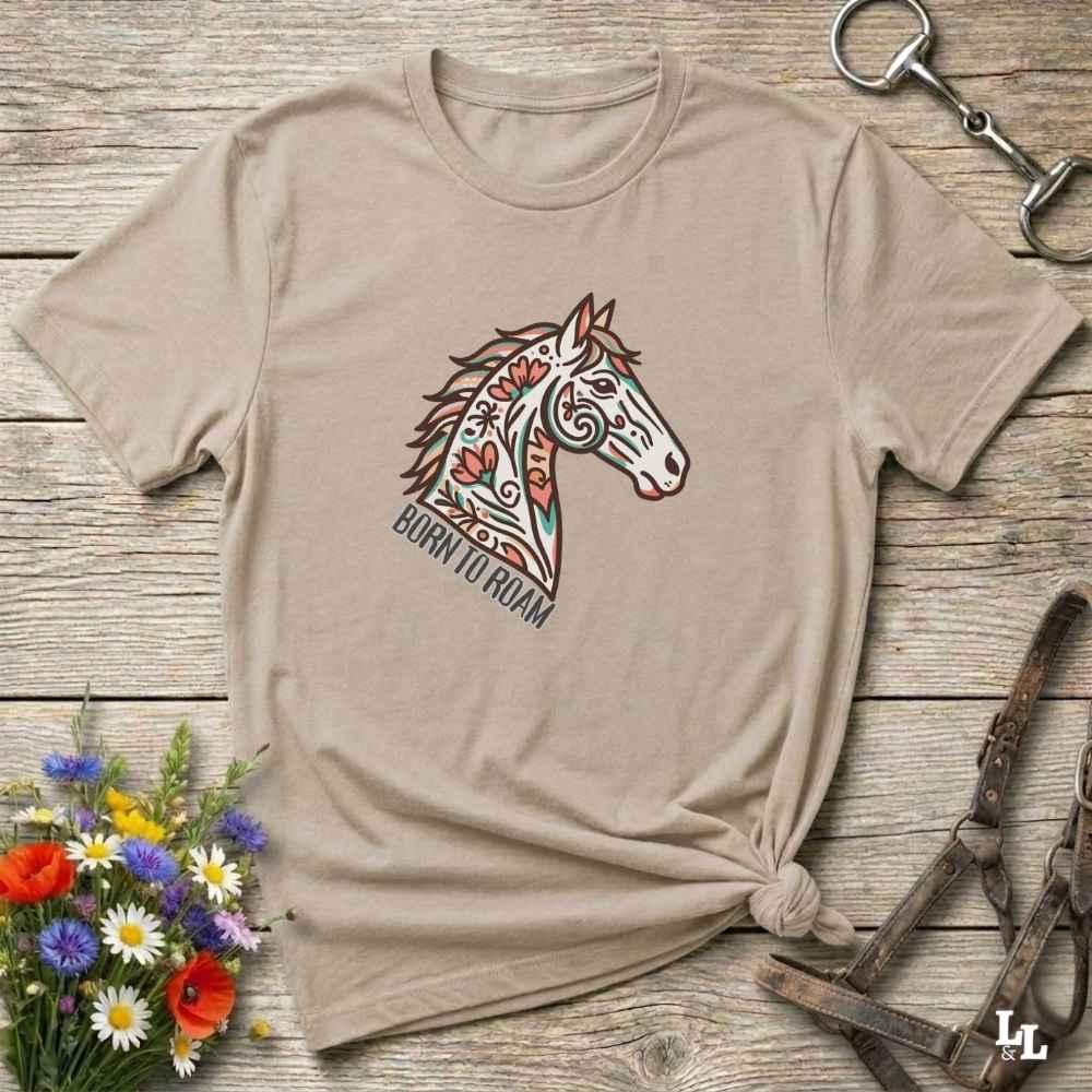 Beige t-shirt with a horse graphic and 'Born to Roam' text on a wooden surface with flowers and equestrian equipment.