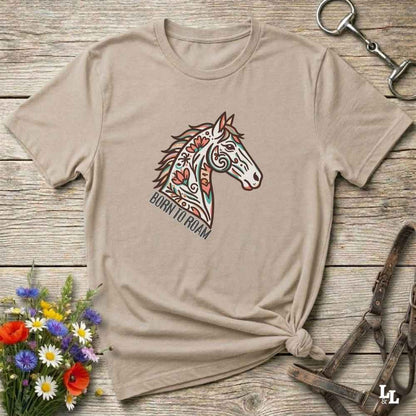 Beige t-shirt with a horse graphic and 'Born to Roam' text on a wooden surface with flowers and equestrian equipment.