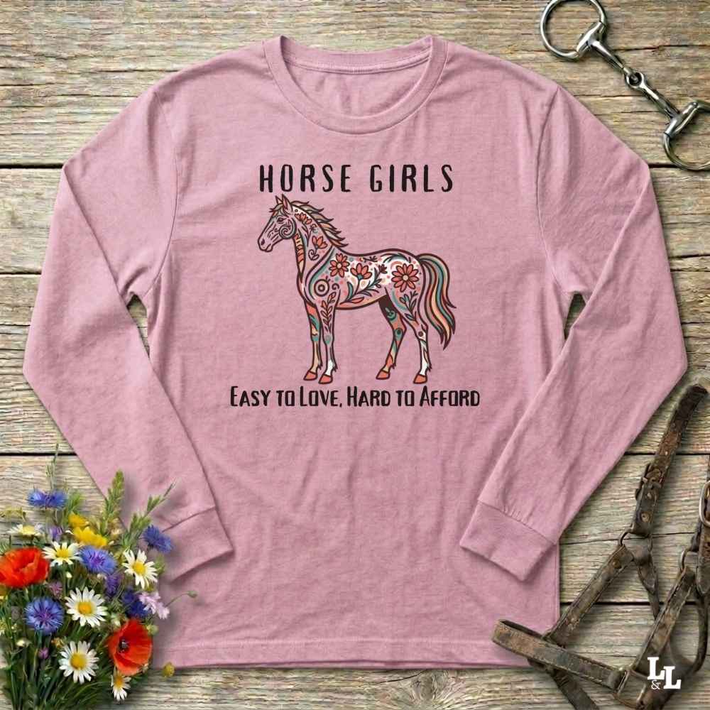 Pink long-sleeve shirt with a horse graphic and text on a wooden surface with floral and equestrian-themed accessories.