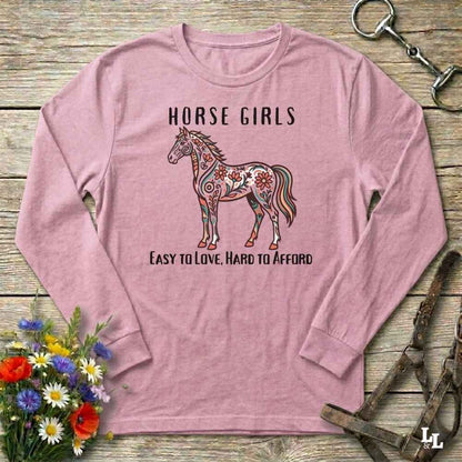 Pink long-sleeve shirt with a horse graphic and text on a wooden surface with floral and equestrian-themed accessories.