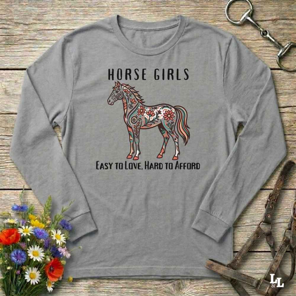 Gray long-sleeve shirt with a horse graphic and text on a wooden surface with flowers and equestrian equipment.