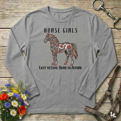Gray long-sleeve shirt with a horse graphic and text on a wooden surface with flowers and equestrian equipment.