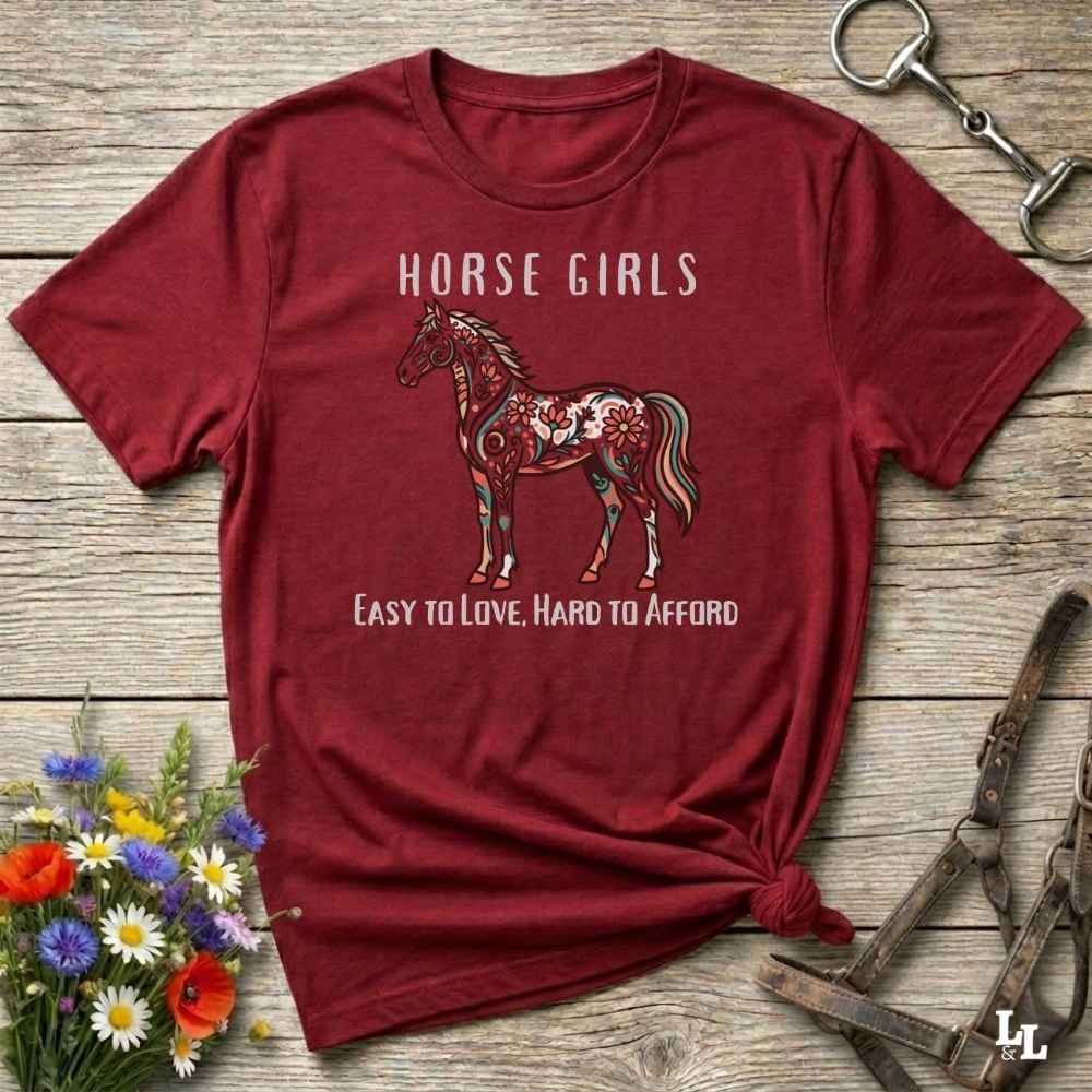 Red t-shirt with horse graphic and text on a wooden surface with floral and equestrian-themed accessories.