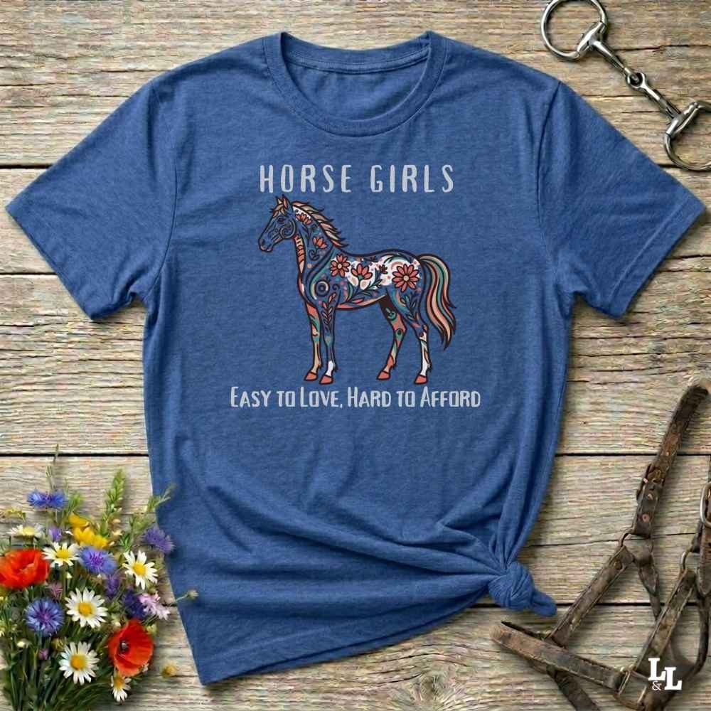 Blue t-shirt with a horse graphic and text on a wooden surface with floral and equestrian accessories.