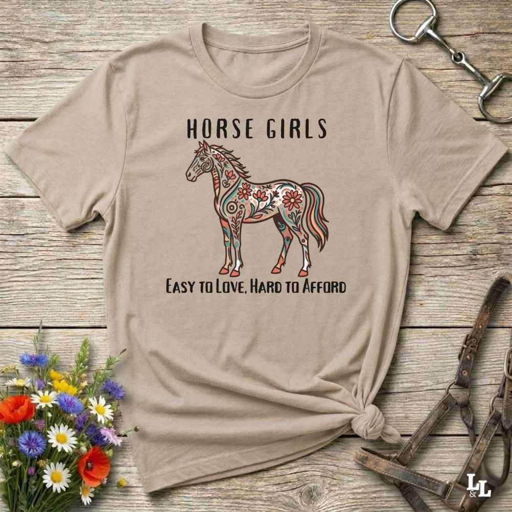 Beige t-shirt with a horse graphic and text on a wooden surface with flowers and equestrian tools.