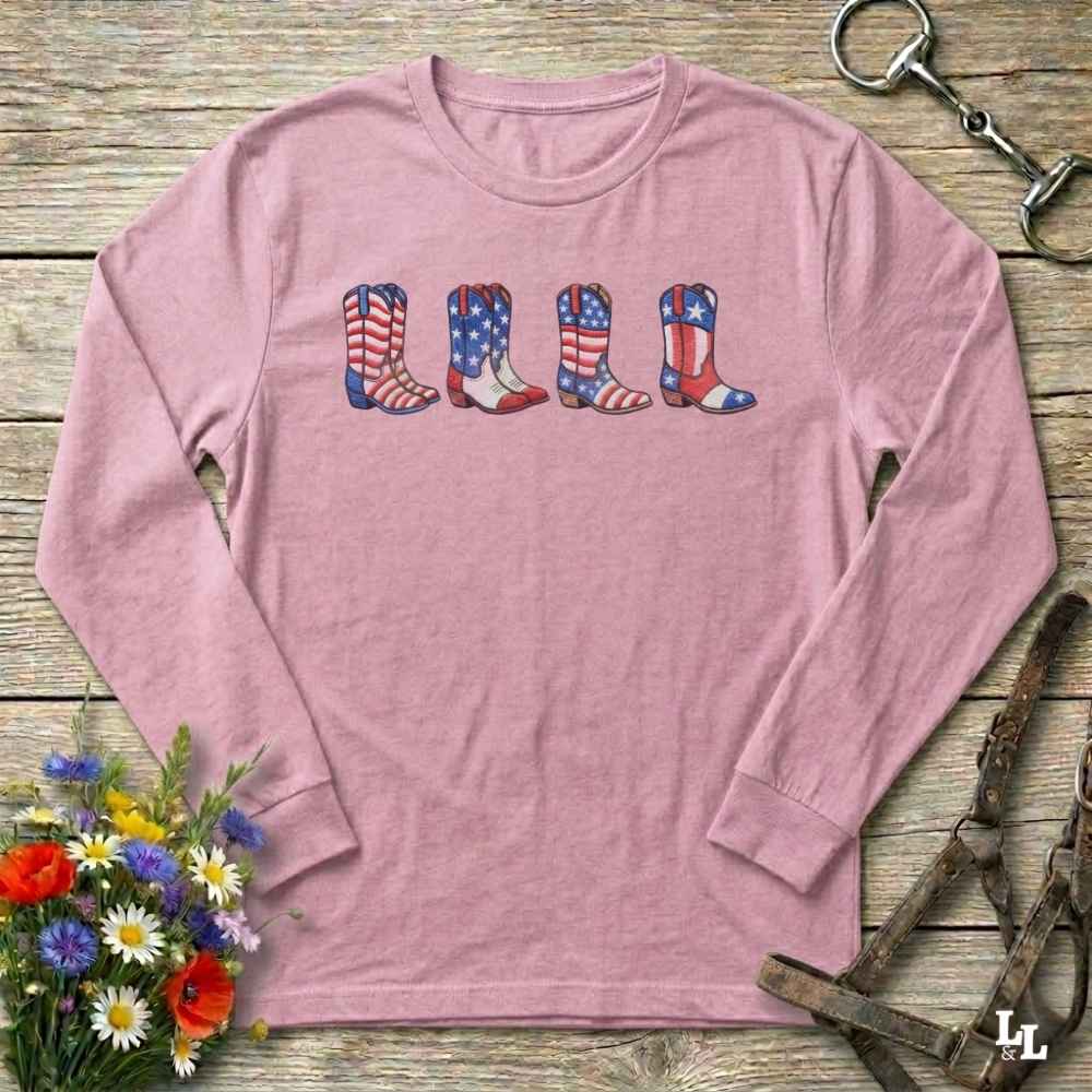 Pink long-sleeve shirt with American flag cowboy boots design on a wooden surface with floral and equestrian elements.