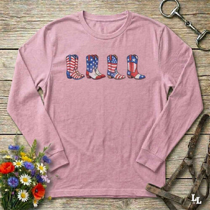 Pink long-sleeve shirt with American flag cowboy boots design on a wooden surface with floral and equestrian elements.