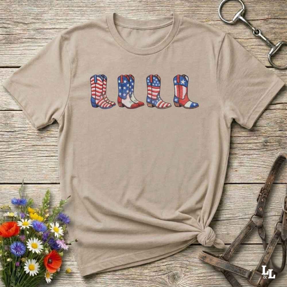 Beige t-shirt with American flag cowboy boots design on a wooden surface with flowers and a horse bit.