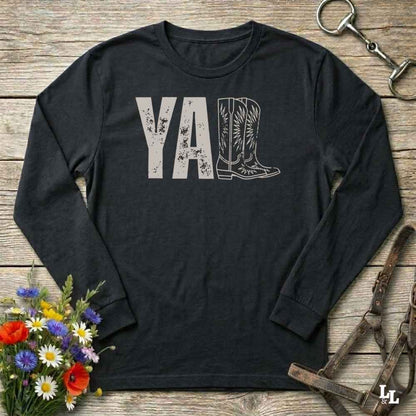 Black long-sleeve shirt with 'YALL' and boot design on a wooden barn floor with flowers and horse equipment.