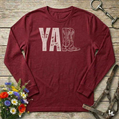 red  long-sleeve shirt with 'YALL' and boot design on a wooden barn floor with flowers and horse equipment.