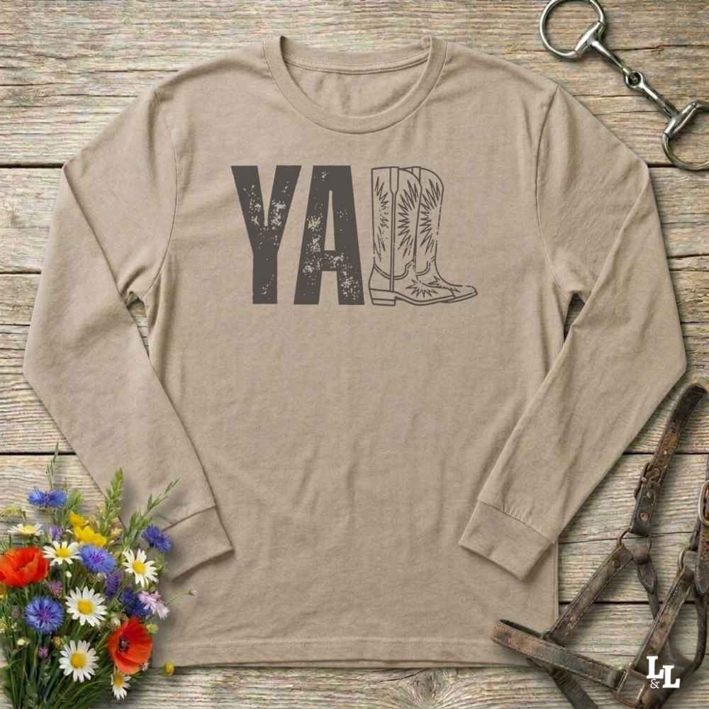 beige  long-sleeve shirt with 'YALL' and boot design on a wooden barn floor with flowers and horse equipment.