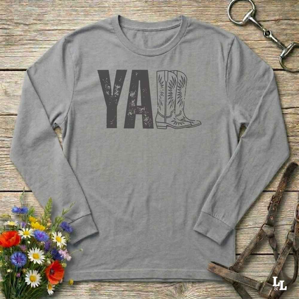 gray  long-sleeve shirt with 'YALL' and boot design on a wooden barn floor with flowers and horse equipment.