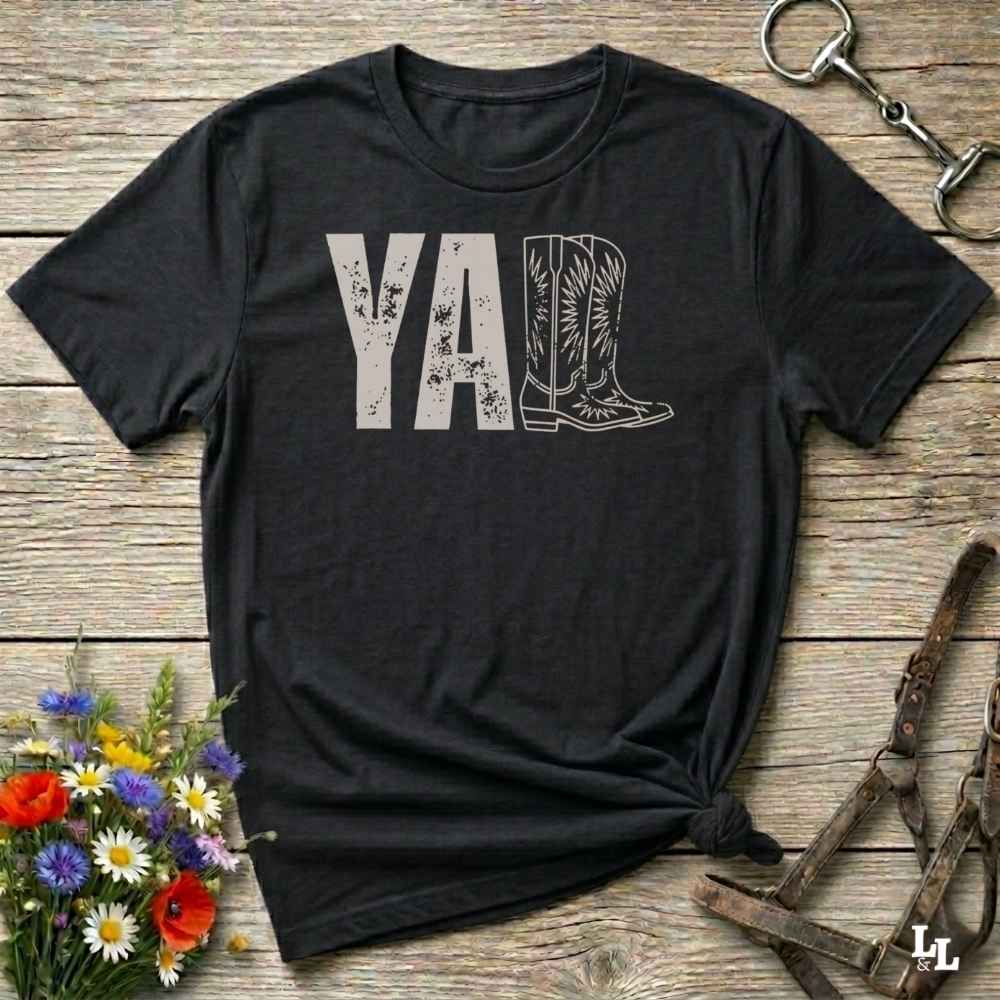 black t-shirt lying flat on wooden barn floor, with graphic design saying "YALL" with the l's being cowboy boots.