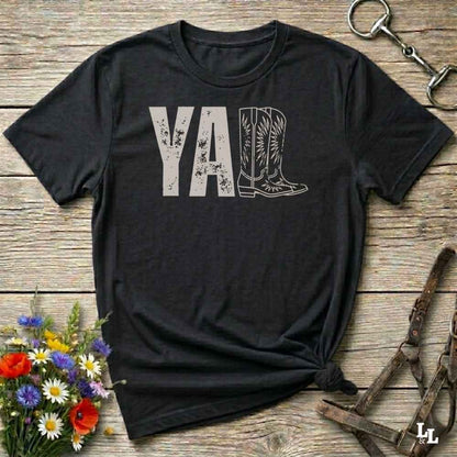 black t-shirt lying flat on wooden barn floor, with graphic design saying "YALL" with the l's being cowboy boots.