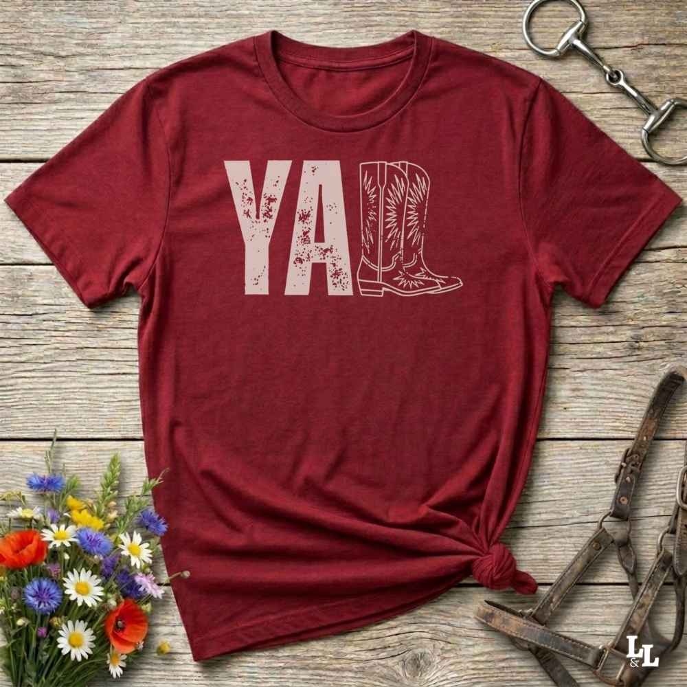 red t-shirt lying flat on wooden barn floor, with graphic design saying "YALL" with the l's being cowboy boots.