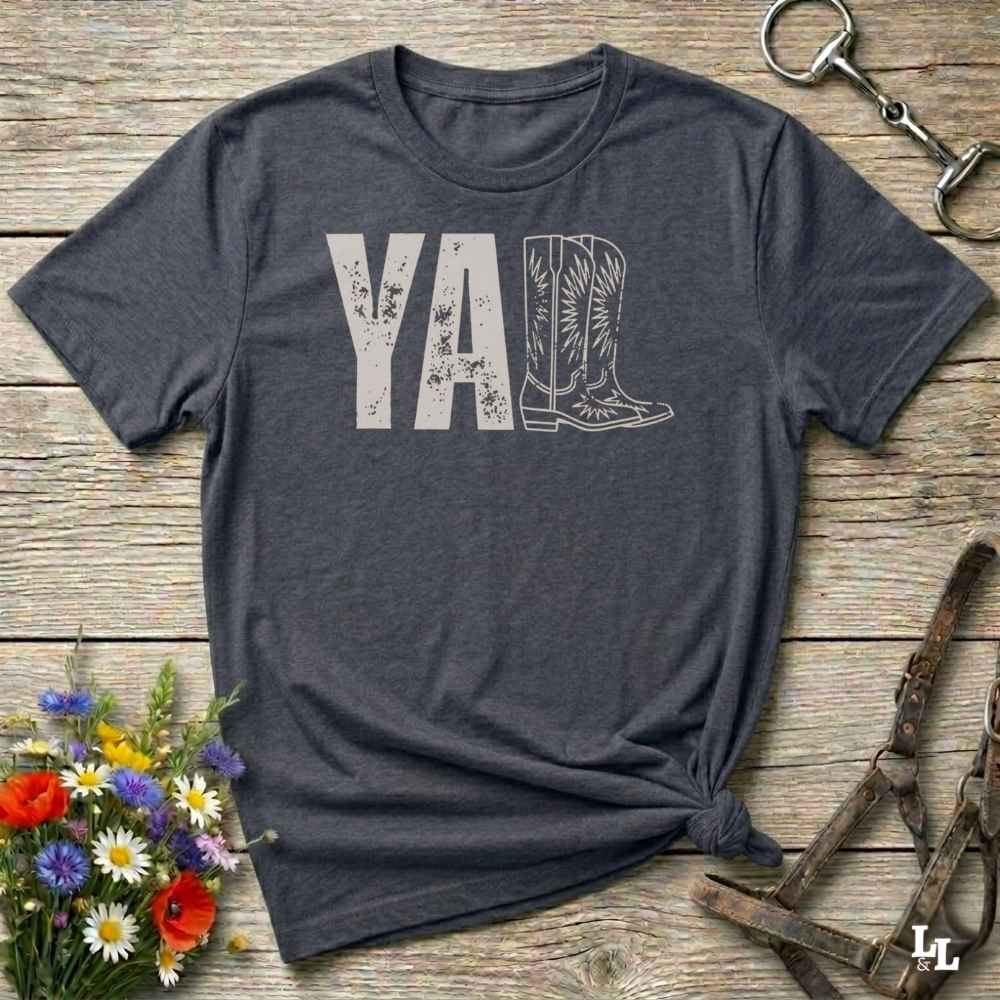 gray t-shirt lying flat on wooden barn floor, with graphic design saying "YALL" with the l's being cowboy boots.