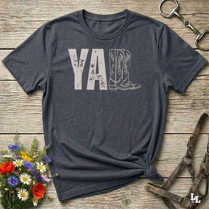 gray t-shirt lying flat on wooden barn floor, with graphic design saying "YALL" with the l's being cowboy boots.