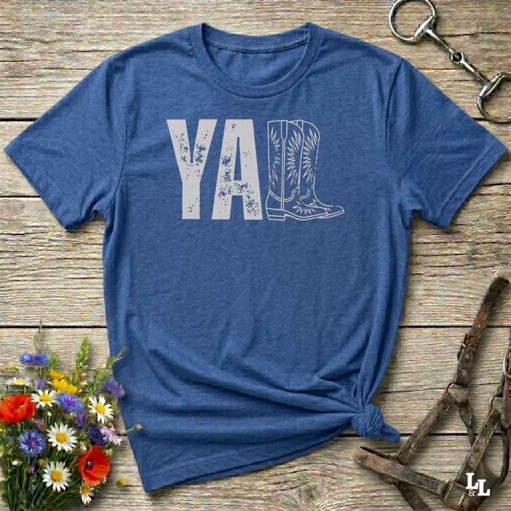 blue t-shirt lying flat on wooden barn floor, with graphic design saying "YALL" with the l's being cowboy boots.