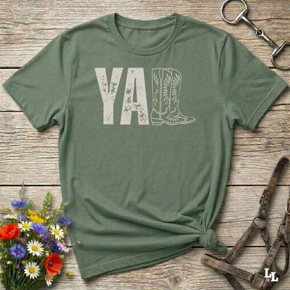green t-shirt lying flat on wooden barn floor, with graphic design saying "YALL" with the l's being cowboy boots.