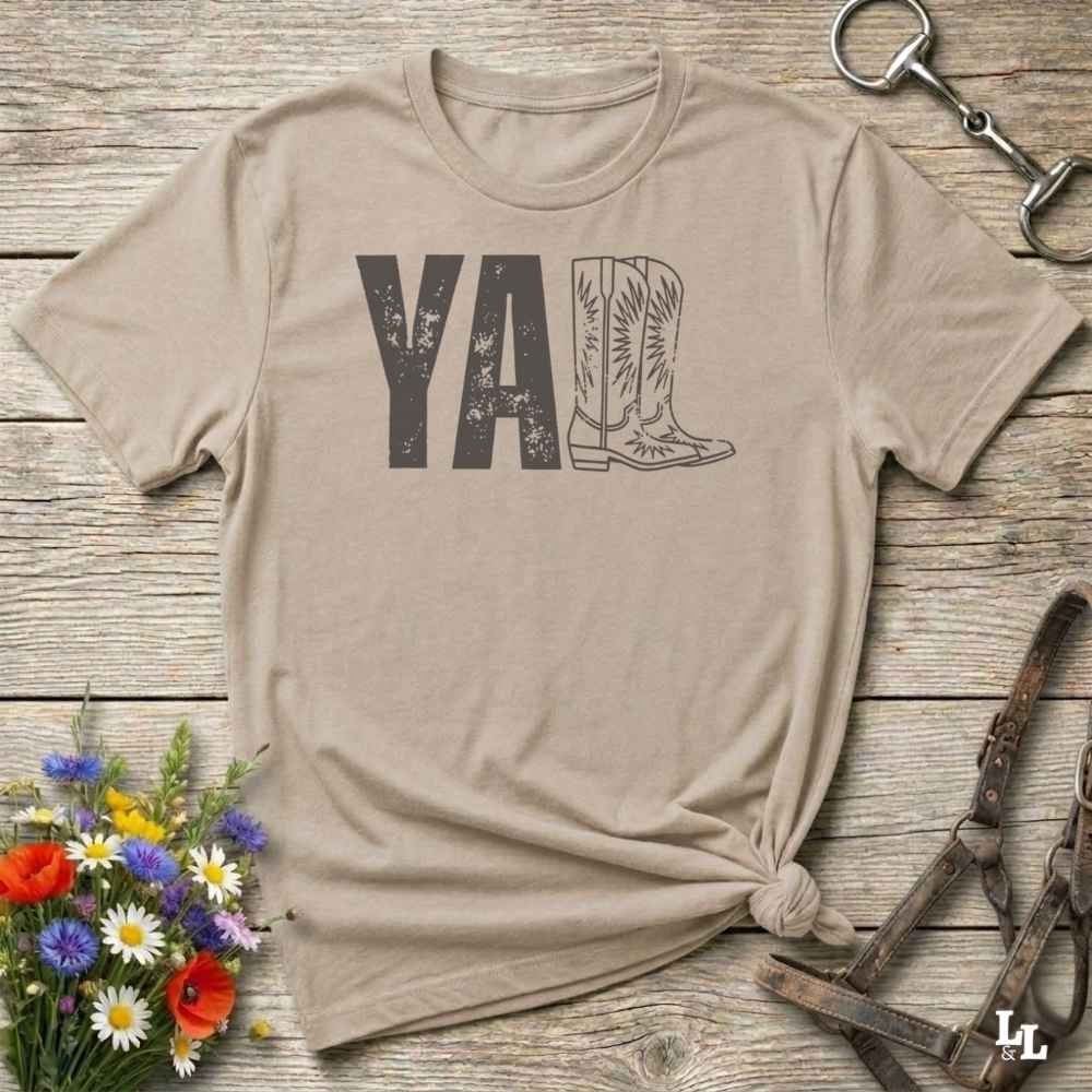 beige t-shirt lying flat on wooden barn floor, with graphic design saying "YALL" with the l's being cowboy boots.