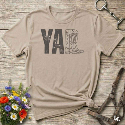 beige t-shirt lying flat on wooden barn floor, with graphic design saying "YALL" with the l's being cowboy boots.