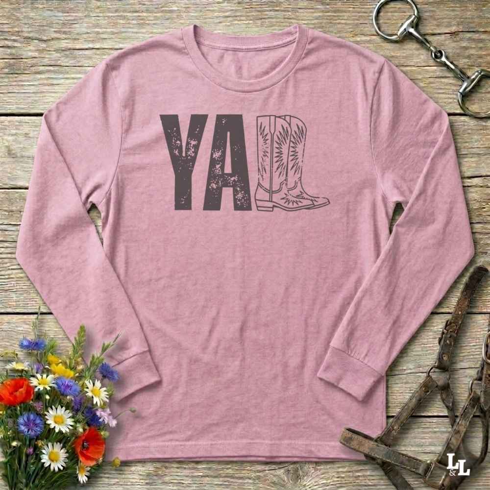 pink  long-sleeve shirt with 'YALL' and boot design on a wooden barn floor with flowers and horse equipment.