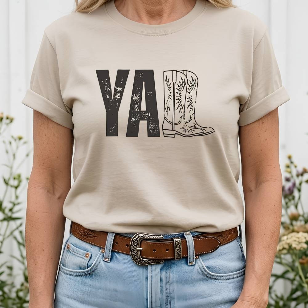 t-shirt lying flat on wooden barn floor, with graphic design saying "YALL" with the l's being cowboy boots.