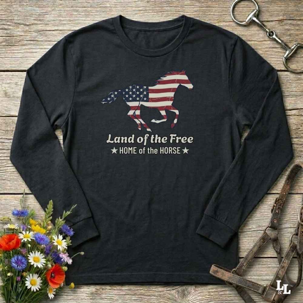 Black long-sleeve shirt with American flag horse design on a wooden surface with flowers and equestrian equipment.