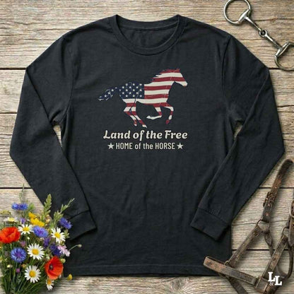 Black long-sleeve shirt with American flag horse design on a wooden surface with flowers and equestrian equipment.