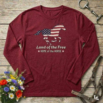Maroon long-sleeve shirt with horse and American flag design on a wooden surface with flowers and equestrian equipment.