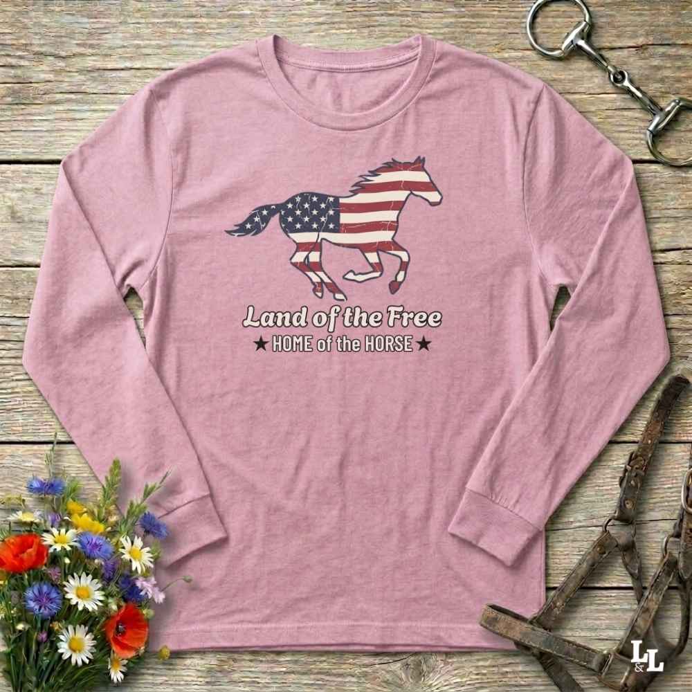 Pink long-sleeve shirt with horse and American flag design on a wooden surface with flowers and horse equipment.