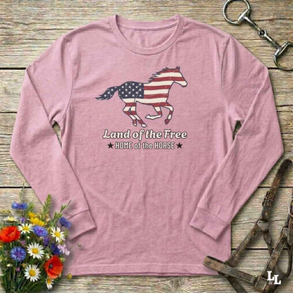 Pink long-sleeve shirt with horse and American flag design on a wooden surface with flowers and horse equipment.